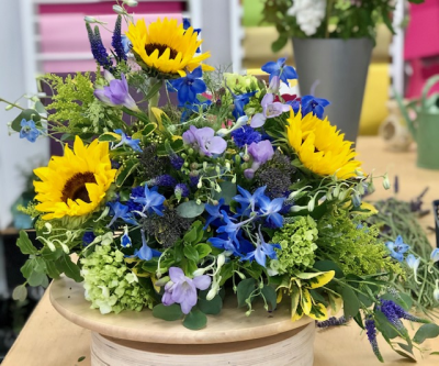 Bright arrangement - Inspired by the joyful hues of a sunlit meadow, this design bursts with the golden cheer of sunflowers, the cool calm of blue delphiniums, and the playful charm of lilac and green