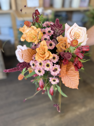 Harvest whisper - A hand-tied burst of countryside charm, this bouquet captures the warmth and richness of early autumn. Blushing peach and sunset orange blooms evoke fields kissed by the golden hour, while the deep reds and burgundy tones bring the richness of ripened berries and turning leaves. Soft pink accents add a gentle nostalgia, like faded quilts and sweet, crisp air. Warm coppery foliage and wild textures complete the look—like a meandering walk through a forest trail, where every leaf crunches underfoot and the light dances between branches. Perfect for a cozy gathering or a rustic celebration.