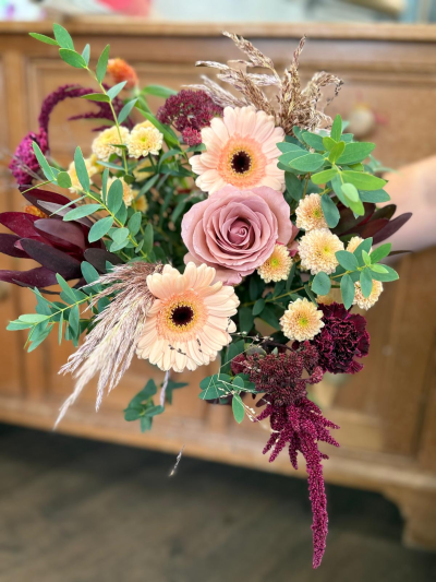 Dusty Meadowlight - Soft and wild, this arrangement whispers of open meadows and sun-dappled fields just before the frost settles in. Muted blush and rose tones mingle with buttery creams and deep berry accents, creating a palette that feels both tender and grounded. Wisps of dried grasses and lush green sprigs bring a sense of movement—like a gentle breeze stirring through the countryside. With its blend of delicate charm and rustic texture, this bouquet carries the soul of a quiet, golden-hour walk through a late-autumn pasture.