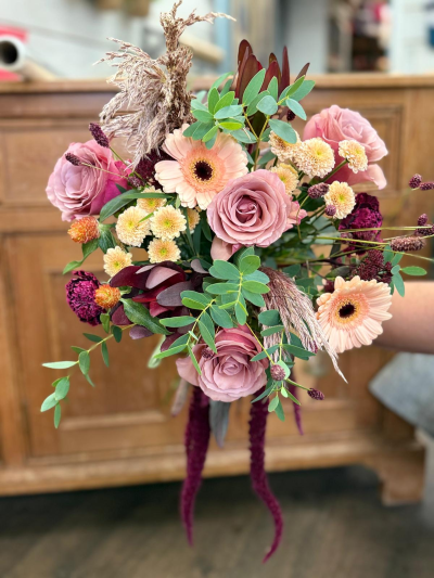 Twilight Prairie - Soft and wild, this arrangement whispers of open meadows and sun-dappled fields just before the frost settles in. Muted blush and rose tones mingle with buttery creams and deep berry accents, creating a palette that feels both tender and grounded. Wisps of dried grasses and lush green sprigs bring a sense of movement—like a gentle breeze stirring through the countryside. With its blend of delicate charm and rustic texture, this bouquet carries the soul of a quiet, golden-hour walk through a late-autumn pasture.