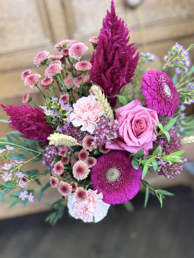 Crimson Glow - This arrangement bursts with the deep warmth of the season—rich berry tones and dusky rose hues flicker like embers against soft, pale highlights. Wisps of feathery textures and spires of velvet-like blooms rise dramatically, while rounded blossoms cluster like tiny harvest moons. Fresh greenery and golden grains weave through it all, giving the bouquet a rustic, fireside charm that captures the cozy glow of late autumn afternoons.