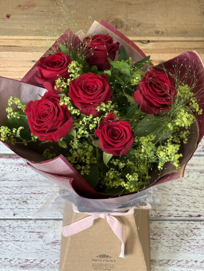 The Sweetheart Six - A breathtaking expression of love, this hand-tied bouquet showcases six luxurious, velvety red blooms—each one a symbol of deep devotion. Their rich, passionate hue is gently framed by delicate wisps of soft green and airy textures that add a touch of enchantment. Cradled in layers of deep burgundy and blush wrapping and finished with a graceful pink satin bow, this bouquet whispers of timeless romance and heartfelt affection—perfect for making any moment feel unforgettable