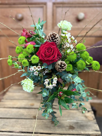 Yuletide Harmony - A classic winter bouquet filled with rich seasonal tones and natural woodland textures. Deep, velvety reds are paired with soft winter greens, while rustic cones and delicate white accents bring a gentle, festive charm. The arrangement is finished with graceful foliage and subtle branches that give it a timeless, handcrafted feel—perfect for adding warmth and elegance throughout the winter season