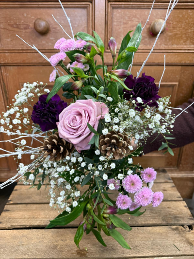 Lavender Snowfall - A soft and dreamy winter arrangement, this bouquet captures the feeling of a quiet snowfall at dusk. Cool lilac tones mingle with deep, velvety shades, creating a tender contrast that feels both elegant and heartfelt. Frost-kissed branches and woodland accents add a touch of magic, while delicate white blossoms give the design a light, airy glow. The result is a romantic winter piece that feels gentle, enchanting, and full of whispered warmth—perfect for gifting love in the colder months.