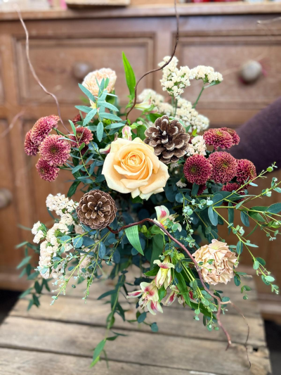 Evergreen whisper - This bouquet captures the quiet beauty of a woodland setting in winter, perfect for adding warmth to your home or gifting to a loved one. Deep berry tones mingle with soft apricot and peach accents, creating a stunning colour palette reminiscent of a glowing fireside hearth.
It features natural, textural elements like winter greenery and real pinecones, giving the design an organic, slightly wild elegance. Finished with delicate filler blooms, this bouquet is a beautiful and heartwarming centrepiece that brings the magic of the forest indoors for the holidays
