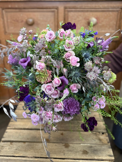 The Snow Queens garden - A sweeping, opulent display of winter romance, this grand arrangement overflows with soft mauves, lingering lilacs, and deep velvet tones. Layers of intricate textures and elegant shapes create a rich tapestry of colour, while shimmering silver accents and woodland details add a festive touch of enchantment. The bouquet feels like a winter garden brought indoors—abundant, lavish, and full of graceful movement. Designed to make a statement, it captures the magic of the season with sophistication and warmth, perfect for celebrating Christmas in true luxury,