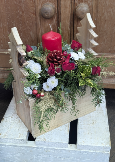 Merry Evergreen arrangement - Bring the warmth of Christmas into any space with this rustic woodland arrangement. A bold red pillar candle rises from a lush bed of fresh evergreens, accented with velvety red rosebuds, crisp white blooms, festive berries, and a glitter-kissed pinecone. The handcrafted wooden crate, topped with carved tree silhouettes, adds a charming chalet-style touch, making it a perfect centerpiece for holiday tables, mantels, or gift-giving. Cozy, cheerful, and beautifully traditional, this arrangement captures the spirit of a magical winter forest.
