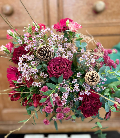 Ruby gift - A charming bouquet brimming with rich holiday colors. It features a stunning center bloom in a deep, magnificent crimson, surrounded by smaller, richly textured flower clusters in shades of ruby red, dark magenta, and deep pink. The composition is beautifully airy, accented by clouds of delicate, misty pink spray flowers that soften the design. Fresh, silvery-green eucalyptus foliage creates a cool backdrop, and the arrangement is finished with rustic pine cones and delicate, bare branches, adding a natural, wintry silhouette. This smaller size still delivers a powerful punch of color and texture, making it an exquisite seasonal gift.