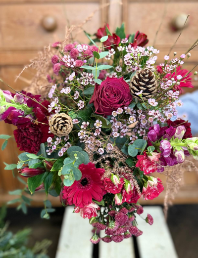 Berry kiss - A dramatic and opulent arrangement celebrating the deepest hues of the holiday season. This bouquet is rich with luxurious, jewel-toned blooms in deep magenta, vibrant ruby red, and striking crimson, complemented by shades of lavender and dark pink for added complexity. It features a central, exquisitely detailed bloom and several smaller, highly-pigmented clusters, giving it a lush, abundant feel. The design incorporates delicate, misty spray flowers in soft pinks that float above the main structure, softening the edges. Natural eucalyptus foliage provides a cool, silvery-green base, and the entire look is finished with rustic pine cones and wisps of dry, bronzed grasses, creating a beautiful depth and a feeling of luxurious winter warmth.