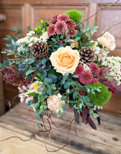 The Gilded forest - This luxurious and expansive arrangement is a true statement piece, celebrating the beauty of the festive season with sophisticated warmth. It showcases an impressive collection of blooms, including striking, plush central petals in creamy apricot and an abundance of smaller, richly toned clusters in shades of deep russet, burgundy, and warm bronze. Delicate highlights of creamy white and pale peach provide elegant contrast. The bouquet is lush with diverse, textured foliage, from silvery-green leaves to dark, dramatic accents, creating a full and enchanting backdrop. Natural elements like rustic pine cones and delicate, curling branches are woven throughout, adding an organic, woodland touch. Unexpected bright green spherical accents and subtle hints of blue-tinged textures add intrigue and depth, making this an exquisite centerpiece for any grand celebration.