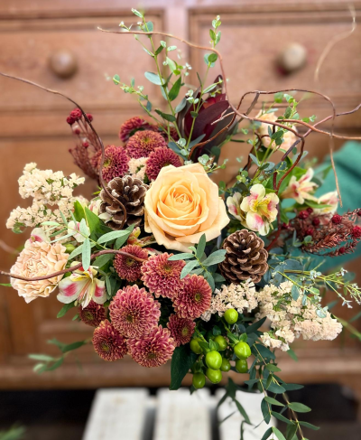 Pine cone dream - An elegant, naturally inspired design featuring a beautiful palette of muted winter tones. The bouquet centers around a large, plush bloom in a soft, creamy apricot color, surrounded by clusters of smaller, russet-toned petals in rich burgundy and bronze shades. Accents of creamy white and pale yellow add a delicate, frosted look. The arrangement is enhanced with generous amounts of mixed foliage, delicate bare branches for height and texture, and unique, bright green berries that offer a pop of unexpected color. The final touch is the inclusion of natural pine cones, grounding the arrangement in rustic, seasonal charm.