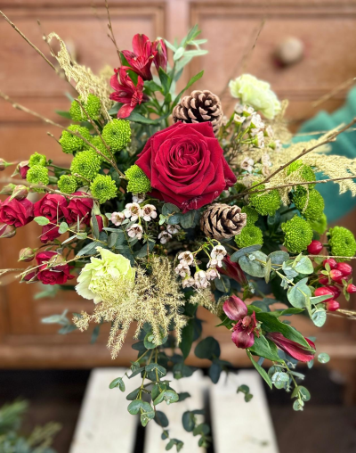 Holly and Hearth - A vibrant and classic Christmas arrangement with a fresh, natural feel. This bouquet is anchored by a magnificent, plush bloom in a rich, signature red, complemented by sprays of smaller red petals and striking, bright lime-green button clusters. The composition is beautifully balanced with accents of creamy white and soft, pale green blooms. Natural textures are key, with long, golden grasses and delicate, bare twigs providing height and movement. Fresh, silvery-green eucalyptus foliage forms a lovely base, and the entire festive look is completed by two perfectly placed natural pine cones, making this a delightful treasure perfect for celebrating the holiday season.
