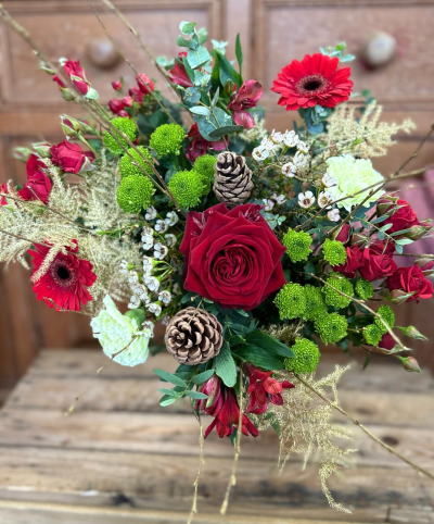 Winter berry luxe - A striking, festive arrangement bursting with the traditional colors of Christmas: deep, velvety reds and vibrant, lime greens. It is expertly gathered with a mix of textures, featuring large, luxurious central blooms and smaller, spray-like accents for depth. The design is complemented by generous wisps of airy, natural foliage and hints of silvery-green leaves, evoking the feel of a winter woodland. Finally, it is perfectly punctuated with natural pine cones that sit nestled amongst the color, adding a beautiful, rustic element and celebrating the warmth and tradition of the holiday season.