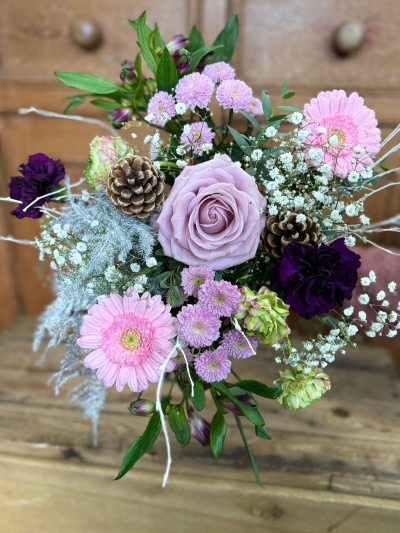 Snowdream - A delightful, compact arrangement that embodies the delicate beauty of a winter morning. This posy features a harmonious mix of cool tones, centered around a soft bloom in dusty mauve or lavender, surrounded by accents of bright pink and deep, striking purple for contrast. The design incorporates beautiful clusters of pale green texture and abundant, airy white filler flowers that evoke a sense of snow and mist. The unique charm of this bouquet comes from the use of soft, silver-toned foliage and subtle white-painted branches, which give the entire arrangement a luxurious, frosted finish. Finished with a single pine cone, this bouquet is an exquisite, sophisticated choice for seasonal gifting.