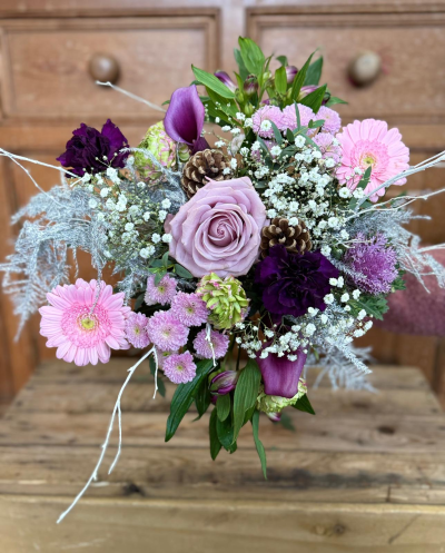 Sugarplum Spell - An enchanting, icy arrangement that captures the serene beauty of a winter's day. This bouquet features a sophisticated blend of cool tones, starring a large central bloom in a soft, dusty lavender, surrounded by clusters of vibrant lilac, deep plum, and delicate, bright pink petals. The design is exquisitely enhanced by abundant, misty white filler flowers and distinctive accents of pale, lime-green texture. What makes this arrangement truly unique is the inclusion of striking, silvery-white foliage and delicate, painted white twigs that evoke the look of frost and ice, transforming it into a luxurious winter dreamscape. Natural pine cones are nestled within, grounding the cool colors with a touch of seasonal texture.