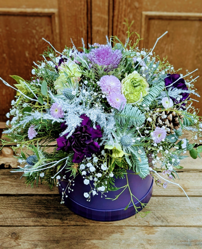 Frost and Plum hatbox - A luxurious hat box arrangement inspired by winter woodlands and festive charm. Soft pastel tones mingle with deep plum accents, creating a gentle contrast that feels both elegant and seasonal. Frosted textures and silvery details give the display a snow-dusted appearance, while touches of natural greenery add warmth and freshness. Subtle berries, pinecones, and delicate filler elements create depth and movement throughout the design. Set in a rich purple hat box, this arrangement brings together winter magic and countryside character—perfect for Christmas gifting or as a show-stopping centrepiece.