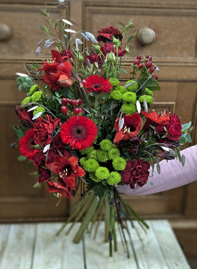 Fireside glow - A tapestry of deep winter colours, this forward-facing bouquet gathers together rich crimson tones and fresh woodland greens, echoing the hush of the countryside as Christmas nears. Textures fold into one another like embers glowing beneath soft snow, while silver-kissed accents catch the light with a quiet shimmer. Full of warmth, depth, and seasonal magic, it brings the feeling of a festive fireside moment into any home.