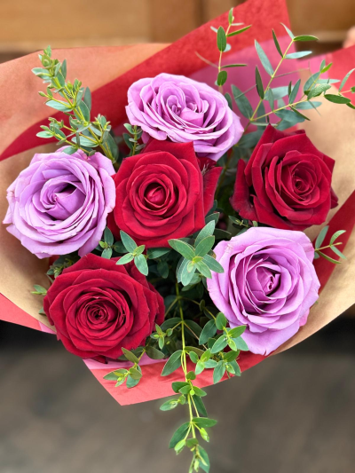 Ever after - A love that speaks in rich contrast and fearless color. Deep crimson unfolds beside soft lavender, each shade intensifying the other in a dance of passion and tenderness. Lush green accents weave through the arrangement like steady breath beneath a racing heart. Wrapped in warm, blushing tones, this bouquet feels intentional and full—romance that doesn’t whisper, but doesn’t need to shout. It’s devotion with depth, desire with balance, and affection that knows exactly where it stands.