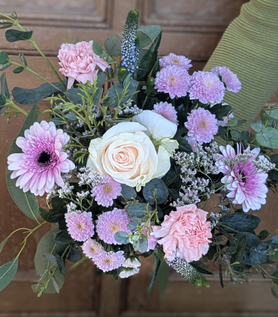 Mother's embrace - A beautifully balanced arrangement in soft blush, pale pink and delicate lilac tones, designed to feel both elegant and heartfelt. At its centre sits a velvety ivory bloom, surrounded by ruffled pastel pink petals and clusters of dainty pom-pom style blossoms. Accents of airy, textured sprigs and fresh, silvery-green foliage add depth and a natural garden feel, while subtle pops of deeper pink create warmth and contrast.

This bouquet has a timeless, graceful look — perfect for celebrating Mum with something that feels thoughtful, feminine and full of love. The mix of textures gives it a hand-tied, just-picked charm while still feeling luxurious and refined.

Please note: stems and varieties may vary slightly depending on seasonal availability, but the overall colour palette, style and value will always be carefully maintained.