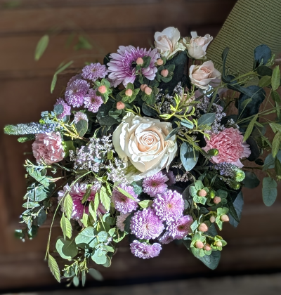 Mother’s Day Garden Glow - A radiant, garden-inspired arrangement in soft blush, pale pink and delicate lilac tones, beautifully balanced with fresh greenery and textural accents. A pale pink statement bloom sits at the heart, surrounded by ruffled pink petals, dainty pom-pom clusters and cheerful daisy-style faces. Sprigs of berry-like detail and airy filler stems weave throughout, adding depth, movement and a naturally gathered feel.  This design feels abundant yet elegant — full of warmth, light and thoughtful detail. Perfect for celebrating Mum with something that feels lovingly hand-tied and effortlessly beautiful.  
Please note: stems and varieties may vary depending on seasonal availability, but the overall colour palette, style and value will always be maintained.